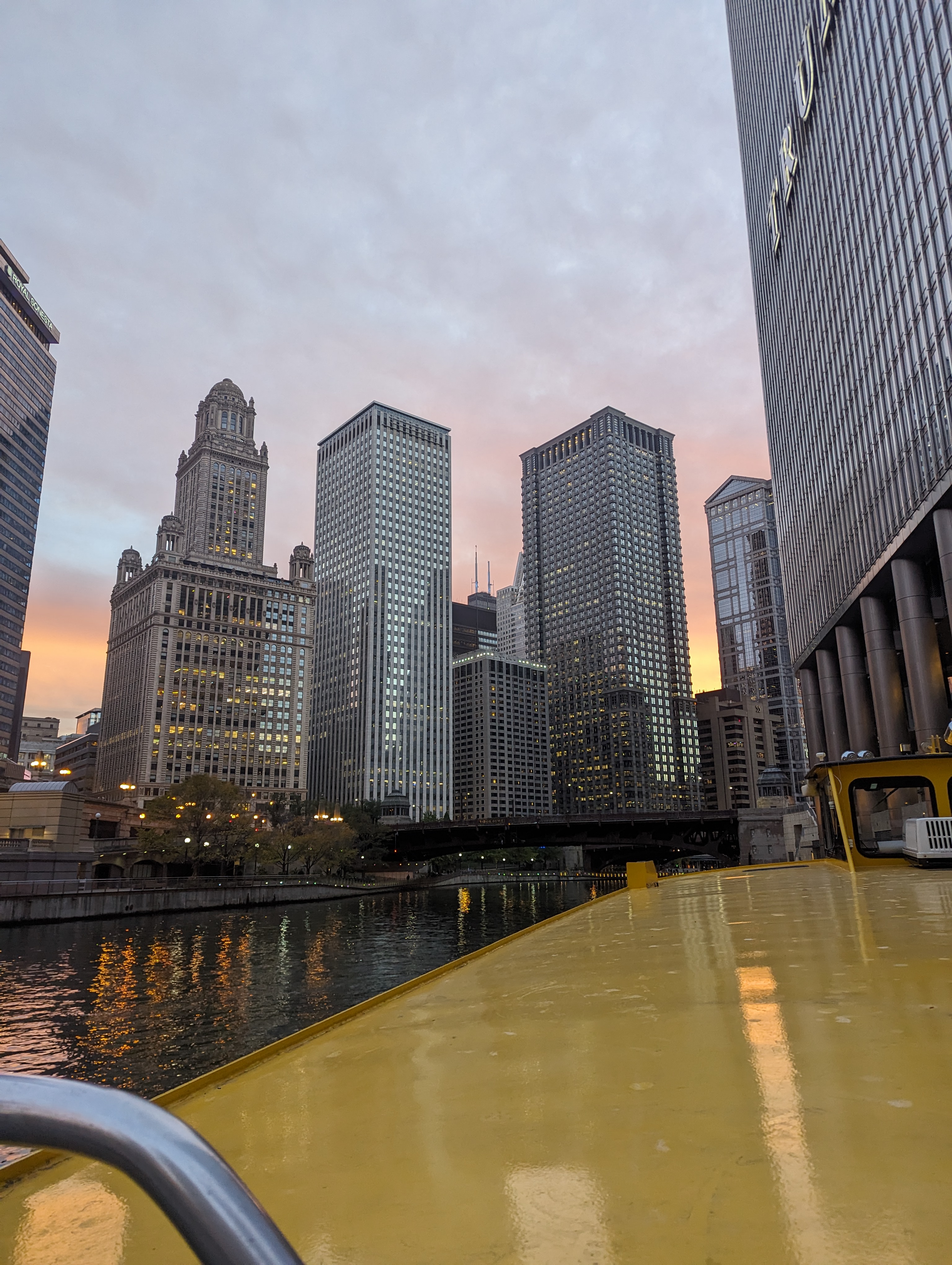 A yellow boat on the Chicago river surrounded by skyscrapers (specifically trump tower to the right and the jewler's building to the left) as the sun sets in the distance.