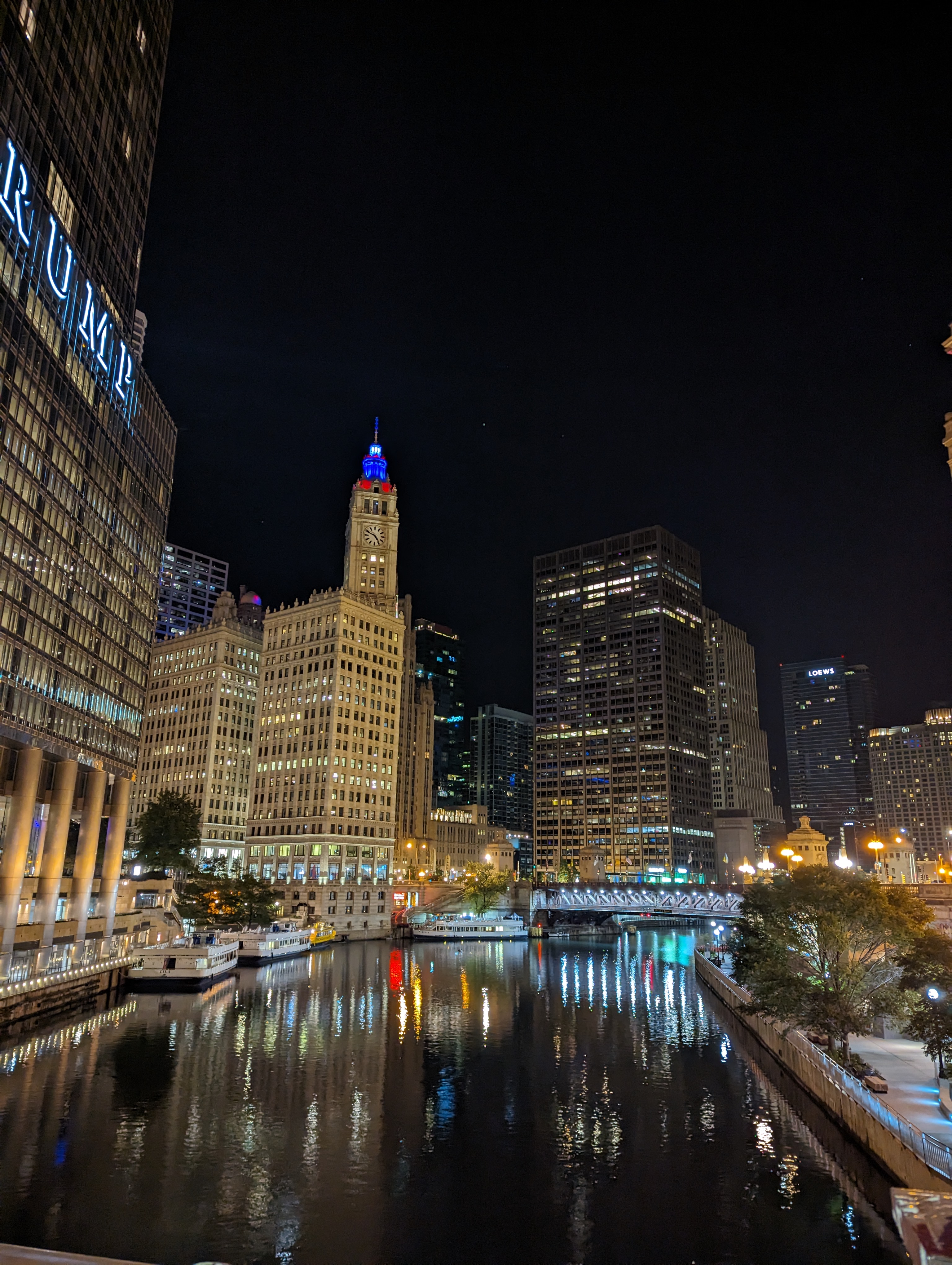 The chicago river as seen from the Wabash street bridge. A riverwalk with trees and benches can be seen from the right. Immediately down the river are Wendella's at trump tower in the wrigley building, with 4 boats being docket. It is nigh time and the top of the Wrigley building is showing blue and red for the colors of the American flag.