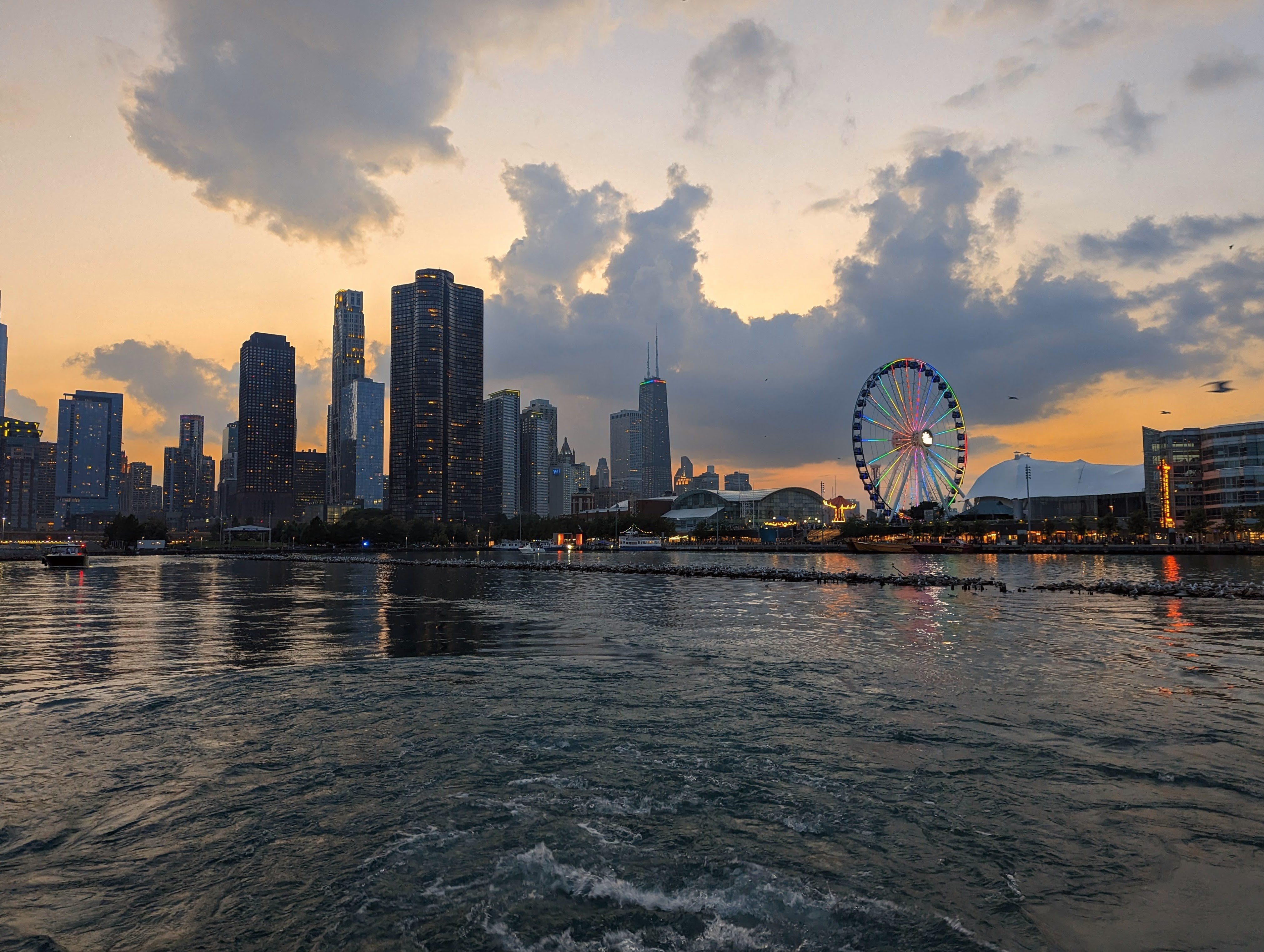 The nothern skyline of Chicago as seen form the Chicago river basin. The John Hancock building and the Navy Piero ferris wheel can be seen. The sunset contains many warm colors, such as orange. All buildings in the shot with lights have the pride colors displayed, as it was taken during pride month.