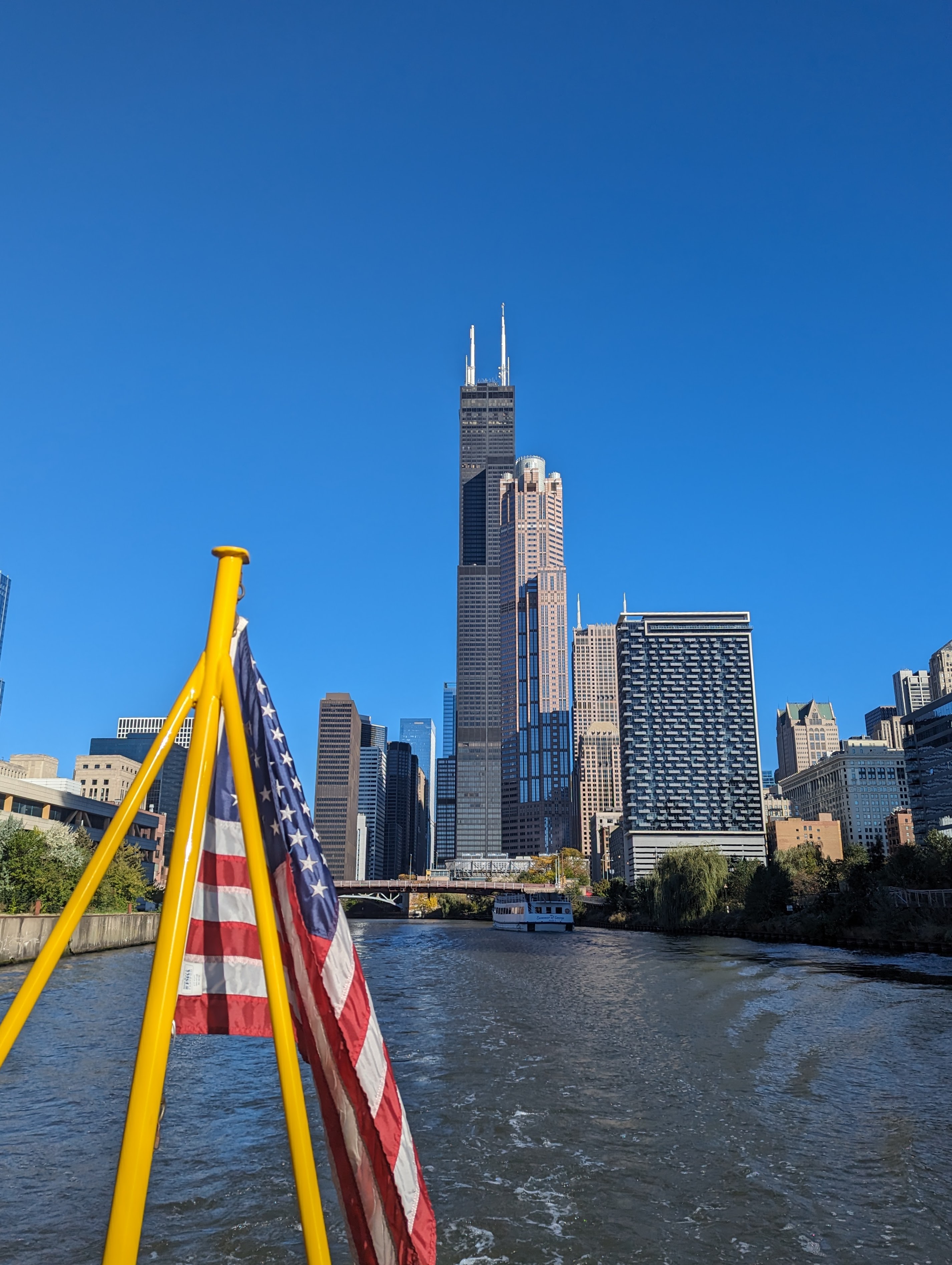 The skyline of Chicago as seen from the south branch of the Chicago river. The sears tower sits front and center. In the foreground is an American flag, as this shot was taken on the back of a boat.